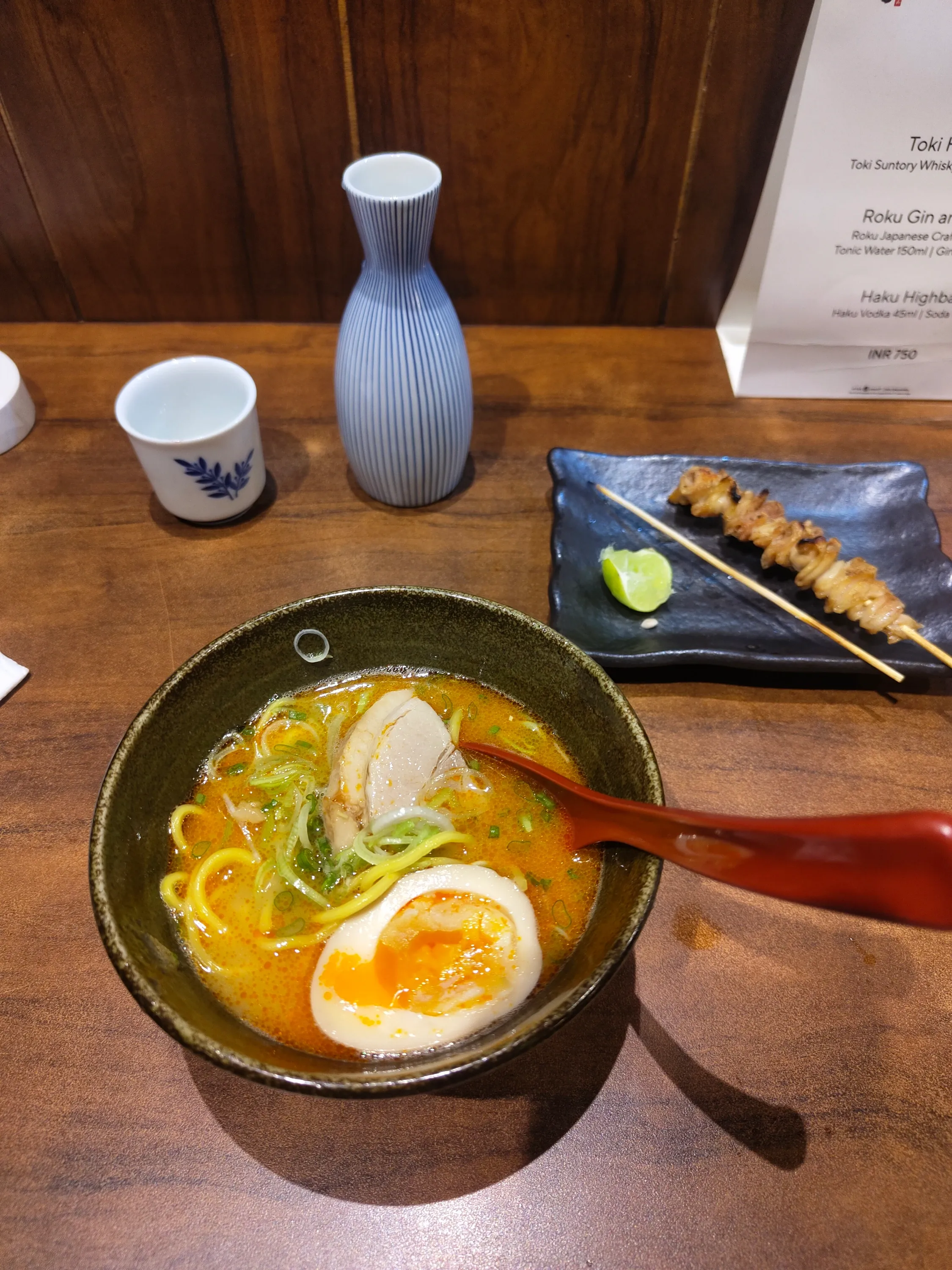A bowl of kuuraku classic ramen, with a half eaten chicken skin yakitori and some plum sake in the background