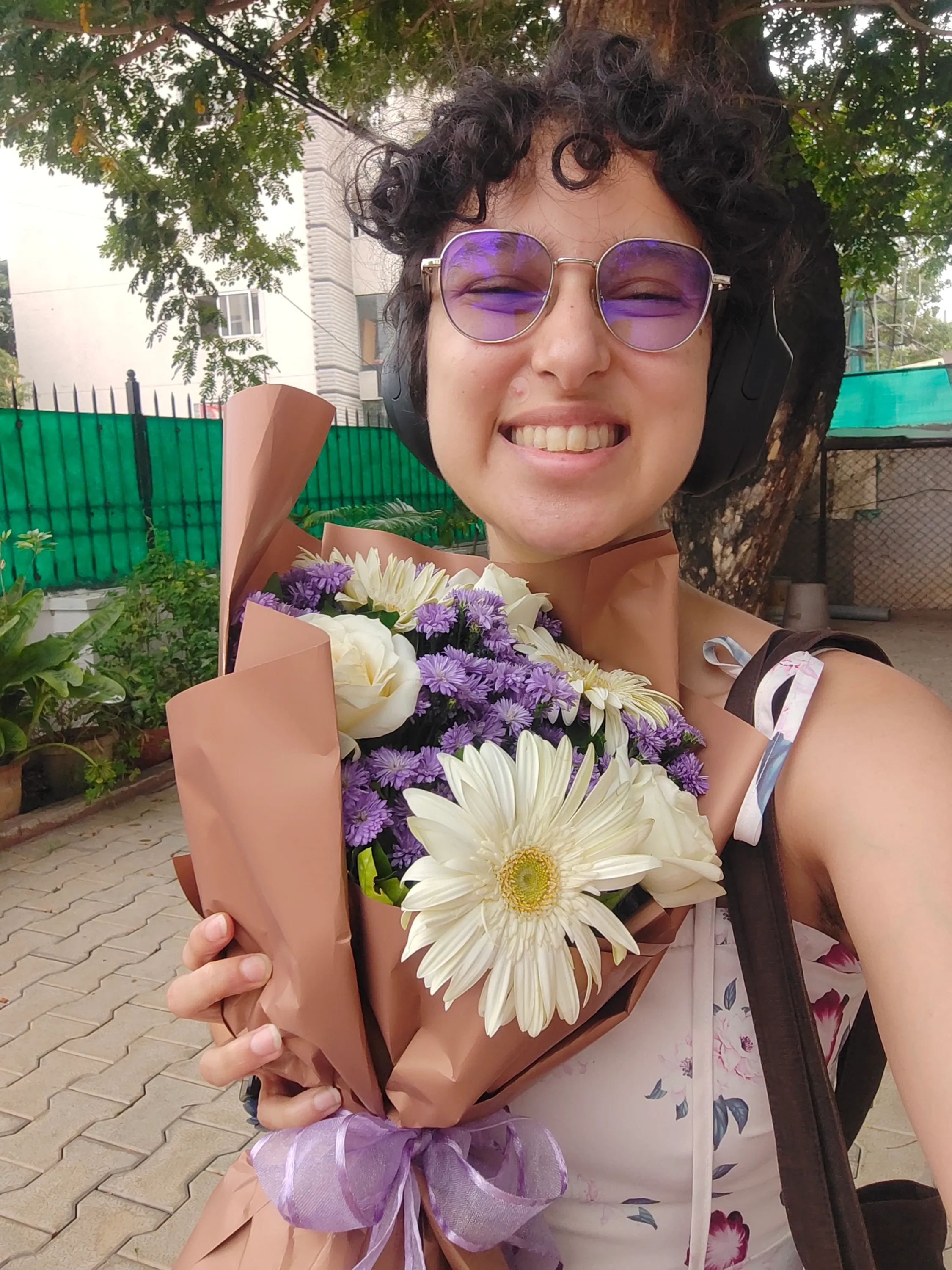 tanvi smiling brightly, the sun reflecting purple from her glasses. She is holding a bouquet of white and purple flowers.